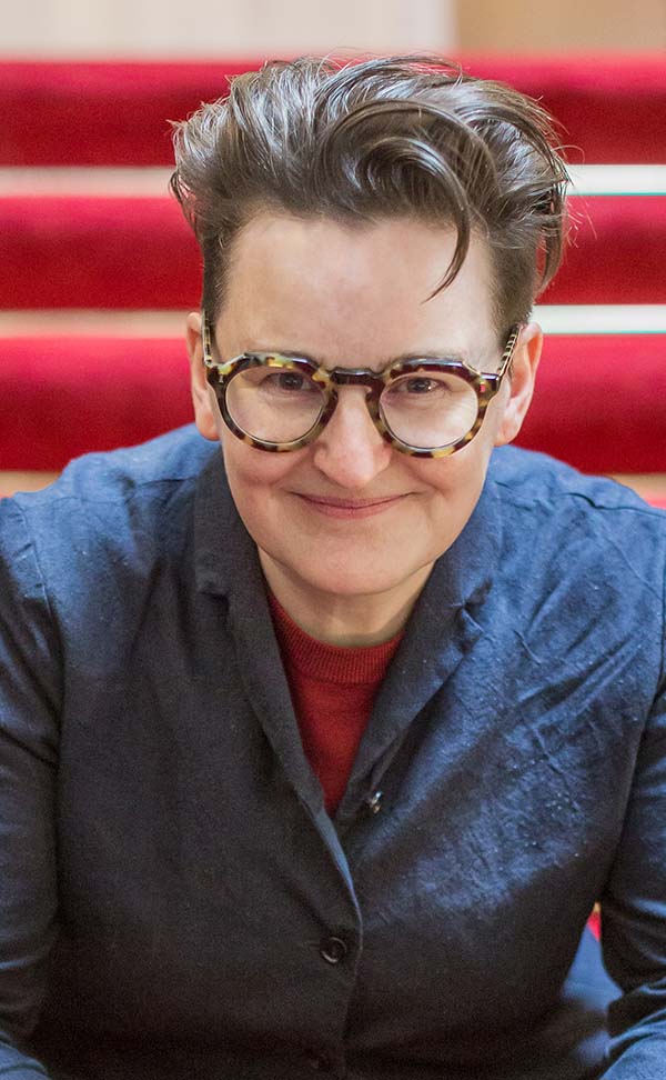 Image of a white woman with short dark hair, wearing a blue shirt and glasses. The woman is smiling.